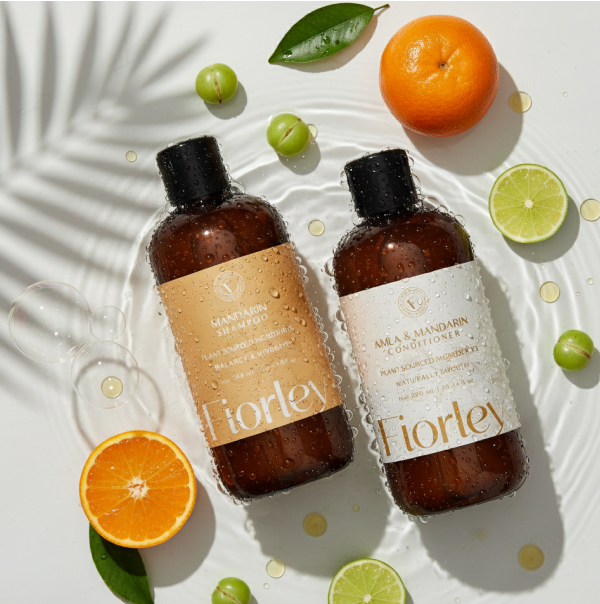 Mandarin shampoo and amla conditioner laying in water with palm leaf shadow, surrounded by amla fruit, leaves and mandarins